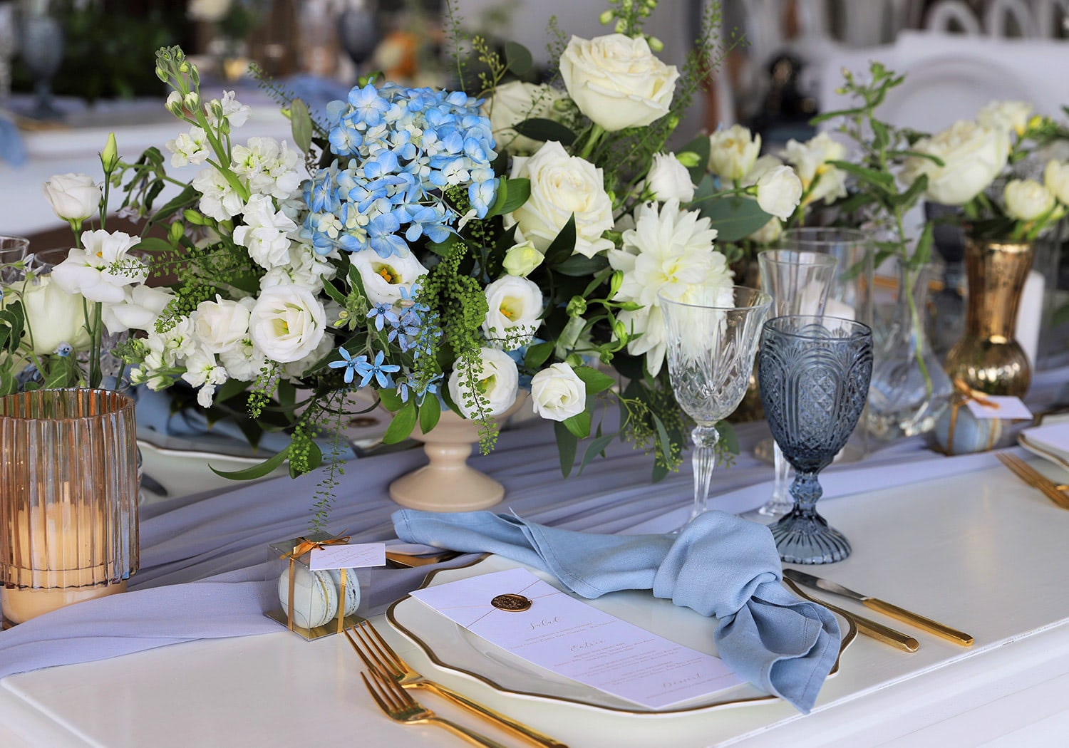Spring table centerpiece with white and blue flowers, roses, and greenery styled for a formal celebration.